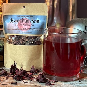 May include: A brown paper pouch labeled Blood Moon Brew containing loose leaf herbal tea, accompanied by a clear glass mug filled with dark red liquid, scattered dried hibiscus petals, and a tall black candle with a lit flame.