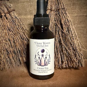 May include: A dark amber glass dropper bottle with a black cap and a white label. The label features the text Three Rivers Herbal Arts, Crone Era, Menopause Support Tincture, and 1 fluid oz./30 ml. The design includes illustrations of moons and bottles.