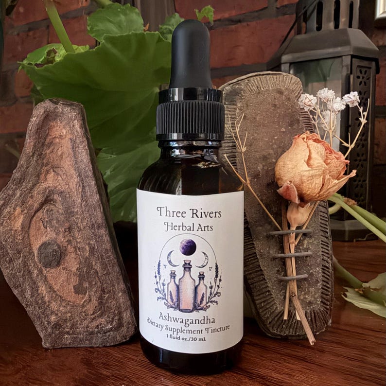 May include: A dark glass bottle of "Ashwagandha Dietary Supplement Tincture" by Three Rivers Herbal Arts. The label features a purple moon and bottle illustration. The bottle contains 1 fluid oz. / 30 ml. A black dropper is attached to the bottle.