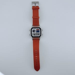 May include: A digital wristwatch featuring a rectangular silver-tone case and a textured orange fabric strap. The watch face displays a digital time of 6:11:48, a small world map graphic, and text including the brand name AWA and 10 YEAR BATTERY.