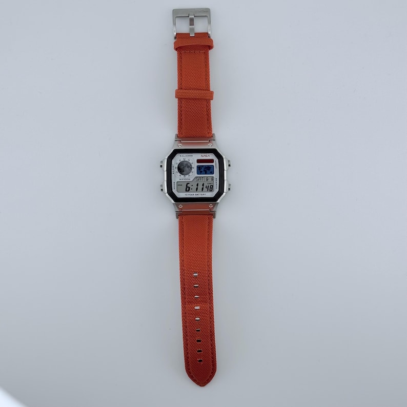 May include: A digital wristwatch featuring a rectangular silver-tone case and a textured orange fabric strap. The watch face displays a digital time of 6:11:48, a small world map graphic, and text including the brand name AWA and 10 YEAR BATTERY.