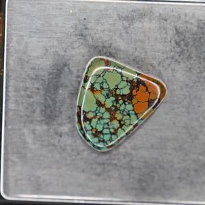 Puede incluir: Una piedra preciosa de turquesa triangular con una matriz compleja de patrones de telaraña en tonos verdes, marrones y negros. La piedra está sobre una báscula digital negra que muestra un peso de 4,69 gramos en la pantalla.