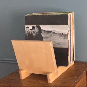 Puede incluir: Un soporte de madera para discos de vinilo, con una parte frontal inclinada y un soporte trasero. Contiene discos de vinilo, con las portadas visibles. La madera es de color marrón claro natural.