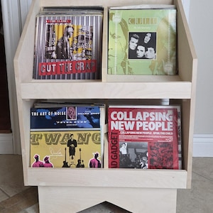 Peut inclure: Un meuble de rangement en bois clair pour disques vinyles, contenant quatre albums. Les pochettes des disques présentent des illustrations colorées et des textes, dont "CUT THE CRAP" et "COLLAPSING NEW PEOPLE". Le meuble a deux étagères et un design angulaire.