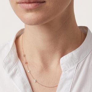Puede incluir: Un collar de oro rosa con las letras B, K y R. El collar se lleva sobre una camisa blanca con cuello. Las letras son pequeñas y espaciadas a lo largo de la delicada cadena. Una joya personalizada.