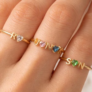 May include: Gold rings featuring letters and heart-shaped gemstones. One ring spells "RUN" with pink and blue gems. Another ring spells "SON" with a green gem. The third ring has the letter "R" and a clear gem.