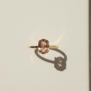 May include: A gold ring featuring a large, oval peach-coloured gemstone. The ring is set on a simple gold band. The ring casts a shadow on a white surface. Jewellery.