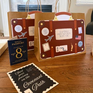Peut inclure: Deux sacs cadeaux en papier brun con&ccedil;us comme des valises vintage avec des autocollants de voyage et le texte BAPTISM PREPARATION SUITCASE. Un livret BAPTISM PASSPORT bleu fonc&eacute; et une carte noire avec le texte Start your journey on the Covenant Path.