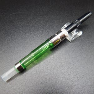 May include: A fountain pen ink converter featuring a transparent green cylindrical reservoir, a silver-toned metal collar, and a black plastic twist mechanism at the top. The bottom end has a clear, translucent plastic nozzle for ink intake.