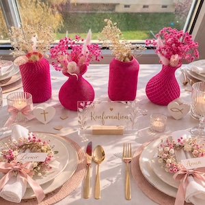 Könnte beinhalten: Ein festlich gedeckter Tisch mit rosa und weißen Blumengestecken, Kerzen und Gedecken. Der Mittelpunkt sind leuchtend pinke Vasen mit Trockenblumen. Jedes Gedeck hat einen Blumenkranz und eine Namenskarte. Ideal für einen besonderen Anlass.