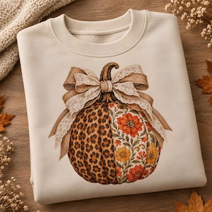 May include: A cream-colored crewneck sweatshirt featuring a central graphic of a pumpkin. The pumpkin is split into two patterns: one side has a brown and black leopard print, and the other has a floral pattern with orange and yellow flowers.
