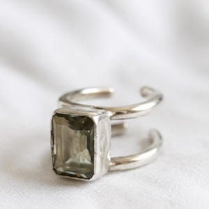 Op de afbeelding: Een zilveren ring met een rechthoekige, olijfgroene edelsteen. De ring heeft een dubbele band, met de edelsteen tussen de twee banden. De ring staat tegen een witte achtergrond.