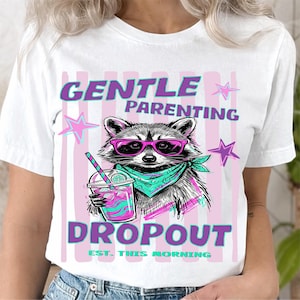 Puede incluir: Camiseta blanca con un gráfico de un mapache con gafas de sol y un pañuelo, sosteniendo una bebida. El texto en la camiseta dice "GENTLE PARENTING DROPOUT EST. THIS MORNING" en morado y turquesa.