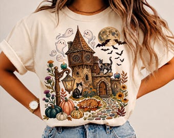 May include: A cream-colored t-shirt featuring a graphic print of a spooky castle, a full moon, bats, and several ghosts. The design includes three cats, various pumpkins, a lit street lamp, and colorful flowers along a stone path.