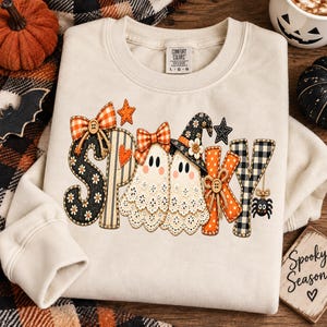 May include: A cream-coloured long-sleeve sweatshirt featuring a graphic print that spells the word SPOOKY. The letters are decorated with patterns including black floral, orange plaid, and black-and-white gingham, with two ghosts, bows, and a spider.