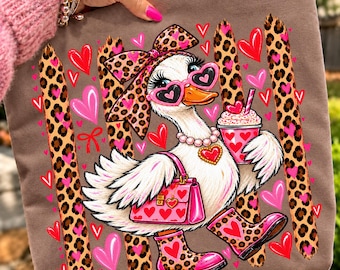 May include: A brown tote bag featuring a graphic of a white duck wearing heart-shaped sunglasses, a pearl necklace, a leopard print bow, and leopard print boots. The duck holds a pink drink and a pink handbag, surrounded by leopard print stripes and hearts.
