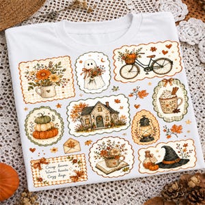 May include: A white t-shirt featuring a collage of autumn-themed illustrations including a house, a ghost, a bicycle with a basket of flowers, pumpkins, a lantern, a witch hat, and a mug, with the text Crisp leaves, Warm hearts, Cozy days.