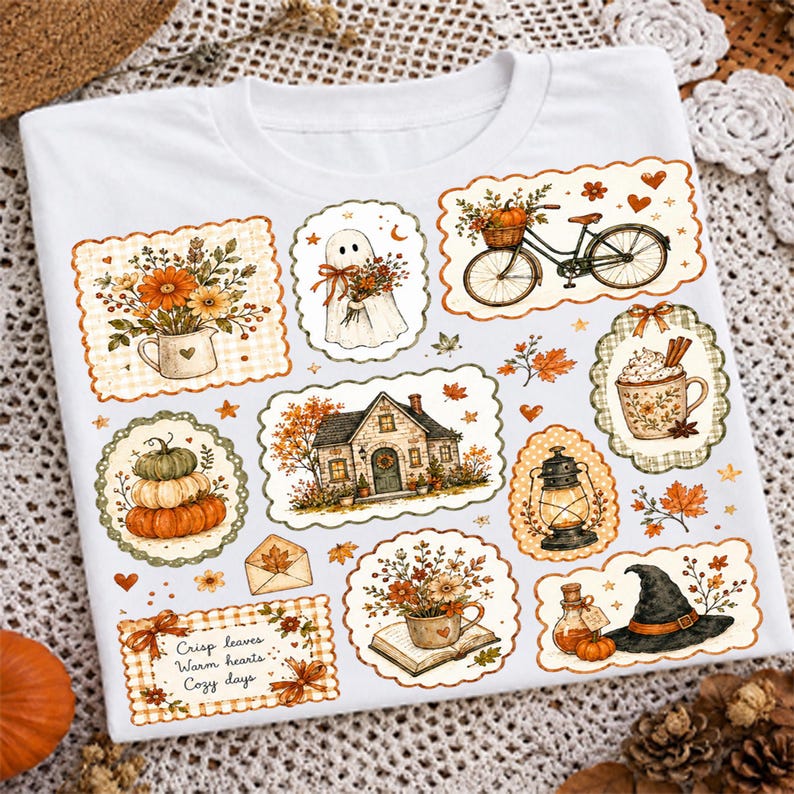 May include: A white t-shirt featuring a collage of autumn-themed illustrations including a house, a ghost, a bicycle with a basket of flowers, pumpkins, a lantern, a witch hat, and a mug, with the text Crisp leaves, Warm hearts, Cozy days.