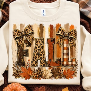 May include: A cream-coloured crewneck sweatshirt featuring the word FALL in large block letters. The letters are decorated with leopard print, brown, and plaid patterns. Two leopard print bows sit atop the letters, surrounded by autumn leaves.