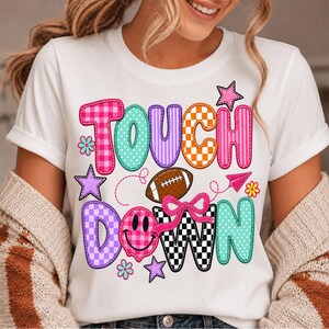 May include: A white t-shirt featuring the text TOUCH DOWN in colorful, patterned block letters. The design includes a brown football, a pink bow, a pink smiley face, stars, flowers, and a small paper airplane graphic printed on the front.