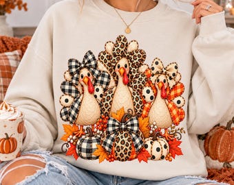 May include: A beige crewneck sweatshirt featuring a graphic of three turkeys adorned with bows. The turkeys are surrounded by pumpkins and autumn leaves. The patterns include black and white buffalo plaid, leopard print, and orange plaid.