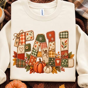 Puede incluir: Una sudadera de cuello redondo color crema con la palabra FALL en letras grandes estilo patchwork. Las letras están decoradas con varios patrones, botones y lazos, y están rodeadas por ilustraciones de calabazas naranjas y blancas y hojas.