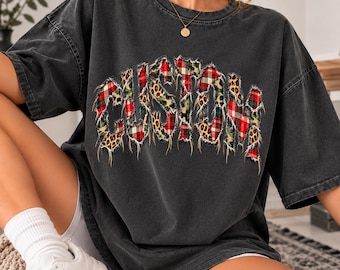 May include: A charcoal grey short-sleeve t-shirt featuring the word CUSTOM in a distressed, frayed-edge font. The letters are filled with a mixed pattern of red and green plaid and brown and black leopard print.