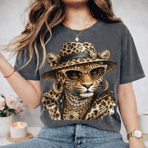 May include: A charcoal gray t-shirt featuring a graphic print of a leopard wearing a leopard-print fedora, gold-rimmed sunglasses, large gold hoop earrings, a layered gold chain necklace, and a black leather-style jacket with gold accents.