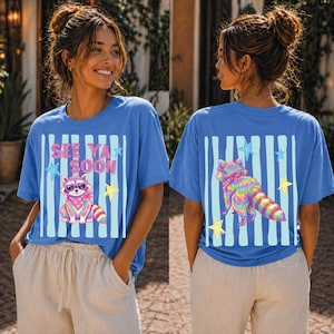 Könnte beinhalten: Blaues T-Shirt mit einem Waschbär-Motiv, das eine Sonnenbrille und ein Bandana trägt. Auf der Vorderseite steht "SEE YA SOON" mit Sternen. Auf der Rückseite befindet sich ein Regenbogen-Waschbär-Design und Sterne. Das T-Shirt hat eine lockere Passform.
