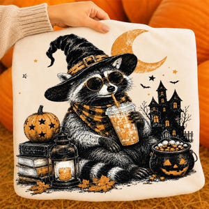 May include: A white square cushion featuring a graphic of a raccoon wearing a witch hat, sunglasses, and a plaid scarf, holding an iced coffee. The design includes a pumpkin, a stack of books, a lantern, a crescent moon, and a haunted house.