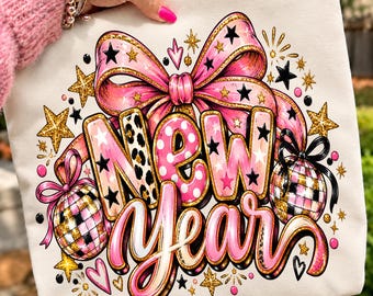 May include: A cream-colored sweatshirt featuring a graphic print that reads New Year in pink, leopard print, and gold-outlined lettering. The design includes a large pink bow, two disco balls, and scattered gold stars and pink hearts.
