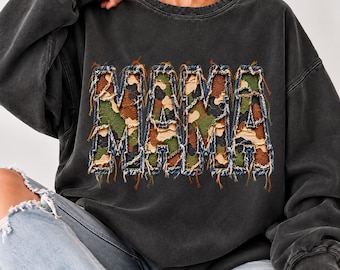 May include: A charcoal gray long-sleeve sweatshirt featuring the word MAMA in large, distressed, camouflage-patterned letters. The letters have frayed denim-like edges with visible stitching, creating a textured, layered appearance.