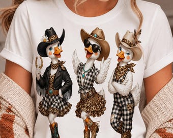 May include: A white t-shirt featuring a graphic of three white ducks dressed in western-style clothing. The ducks wear cowboy hats, leopard print scarves, plaid skirts or overalls, and boots. One duck wears a black fringed jacket and holds a horseshoe.