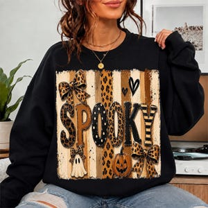 May include: A black long-sleeve sweatshirt featuring a central graphic with the word SPOOKY in large letters. The design includes leopard print patterns, vertical stripes, two bows, a small white ghost, a jack-o'-lantern, and two black hearts.