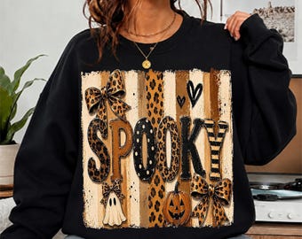 May include: A black long-sleeve sweatshirt featuring a central graphic with the word SPOOKY in large letters. The design includes leopard print patterns, vertical stripes, two bows, a small white ghost, a jack-o'-lantern, and two black hearts.