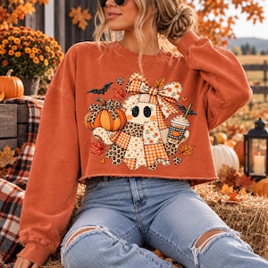 May include: A burnt orange long-sleeve cropped sweatshirt featuring a graphic print of a ghost wearing a large plaid bow. The ghost is surrounded by a pumpkin, a coffee cup, small bats, and autumn leaves in shades of orange, brown, and cream.