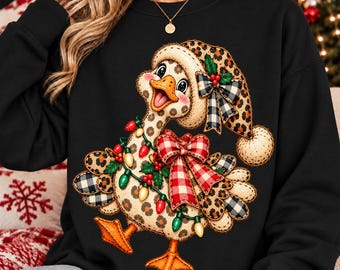 May include: A black long-sleeve sweatshirt featuring a graphic of a duck wearing a leopard print hat with a plaid bow. The duck is decorated with a string of colorful Christmas lights, a large red and white checkered bow, and holly berries.