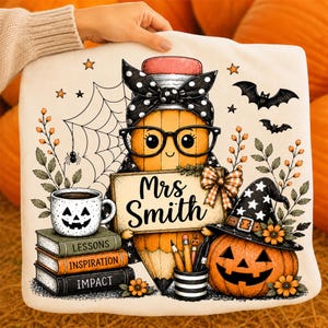 May include: A white square cushion featuring a cartoon pencil character with glasses and a polka-dot bow, holding a sign that reads Mrs Smith. The design includes a jack-o'-lantern, a stack of books labelled Lessons, Inspiration, and Impact, a spider web, and bats.