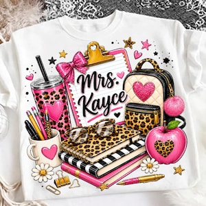 Può includere: Una t-shirt bianca con una stampa grafica raffigurante una cartellina con il testo Mrs. Kayce, un bicchiere con stampa leopardata, uno zaino, una tazza con penne, quaderni, occhiali, una mela rosa e varie stelle, cuori e fiori decorativi.