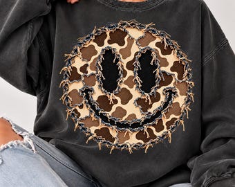 May include: A charcoal gray crewneck sweatshirt featuring a large, circular smiley face appliqué. The smiley face has a brown and cream cow print pattern with frayed denim edges, black oval eyes, and a black curved mouth.