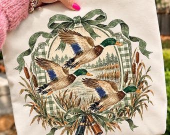 May include: A cream-colored sweatshirt featuring a graphic print of three mallard ducks in flight over a lake. The design includes a green gingham border, a decorative green ribbon bow, cattails, and a pair of binoculars at the bottom.