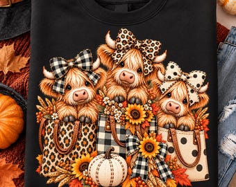 May include: A black sweatshirt featuring a graphic of three fluffy Highland cows, each wearing a patterned bow, sitting inside individual tote bags. The design includes sunflowers, autumn leaves, and a white pumpkin at the base.