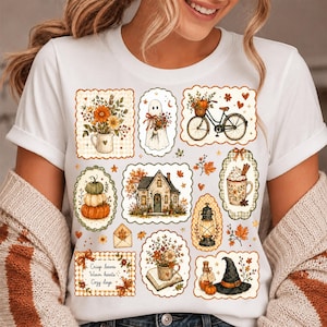 May include: A white t-shirt featuring a collage of autumn-themed illustrations including a ghost, a bicycle with a pumpkin basket, a house, a lantern, a witch hat, and floral arrangements. The text reads, "Crisp leaves, Warm hearts, Cozy days."