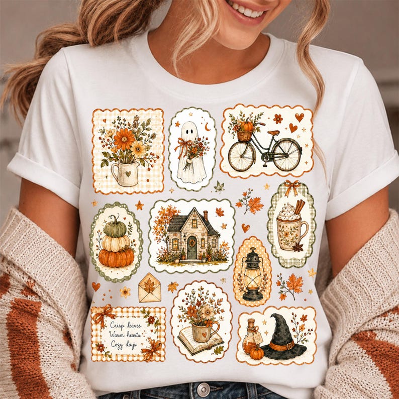 May include: A white t-shirt featuring a collage of autumn-themed illustrations including a ghost, a bicycle with a pumpkin basket, a house, a lantern, a witch hat, and floral arrangements. The text reads, "Crisp leaves, Warm hearts, Cozy days."