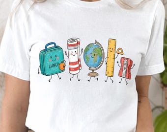 May include: A white short-sleeved t-shirt featuring a colorful graphic print of five anthropomorphic school supplies: a teal lunch box with the word LUNCH, a striped towel, a globe, a yellow ruler, and a red pencil sharpener with a protractor.