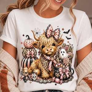 May include: A white t-shirt featuring a graphic of a fluffy highland cow wearing a pink leopard print bow and bandana. The design includes stacked pumpkins, a ghost, a cauldron, a skull, bats, and spiderwebs in shades of pink, black, and gold.