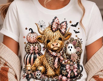 May include: A white t-shirt featuring a graphic of a fluffy highland cow wearing a pink leopard print bow and bandana. The design includes stacked pumpkins, a ghost, a cauldron, a skull, bats, and spiderwebs in shades of pink, black, and gold.