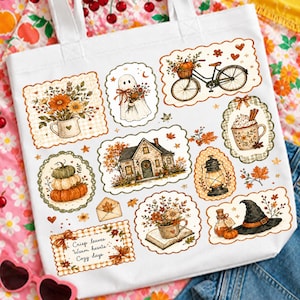 May include: A white canvas tote bag featuring various autumn-themed illustrations including a bicycle with a pumpkin basket, a ghost, a house, a lantern, a witch hat, and floral arrangements. The text reads, "Crisp leaves, Warm hearts, Cozy days."