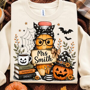 May include: A cream-coloured crewneck sweatshirt featuring a graphic of a cartoon pencil wearing glasses and a polka-dot headband. The pencil holds a sign reading Mrs. Smith, next to a stack of books labelled Lessons, Inspiration, and Impact.