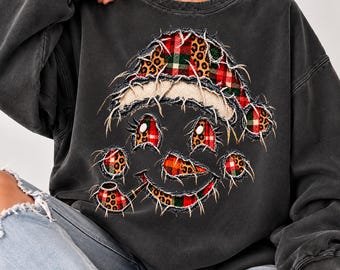 May include: A charcoal gray crewneck sweatshirt featuring a distressed-style graphic of a snowman wearing a Santa hat. The snowman design is composed of frayed-edge patches in red and green plaid and leopard print patterns.